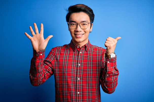 Jeune bel homme chinois portant chemise décontractée et lunettes sur fond bleu montrant et pointant vers le haut avec les doigts numéro six tout en souriant confiant et heureux
. - Photo, image