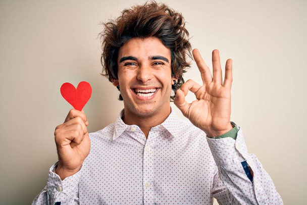 Jeune bel homme tenant le coeur en papier debout sur fond blanc isolé faisant signe ok avec les doigts, excellent symbole
 - Photo, image