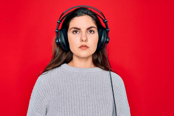 Fiatal gyönyörű lány visel modern fejhallgató zenét hallgatni piros háttér komoly kifejezés az arcon. Egyszerű és természetes a kamerába nézni.. - Fotó, kép