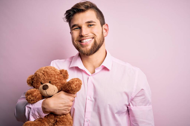 Молодой блондин с бородой и голубыми глазами держит игрушку плюшевого медведя на розовом фоне с счастливым лицом стоя и улыбаясь с уверенной улыбкой показывая зубы
 - Фото, изображение