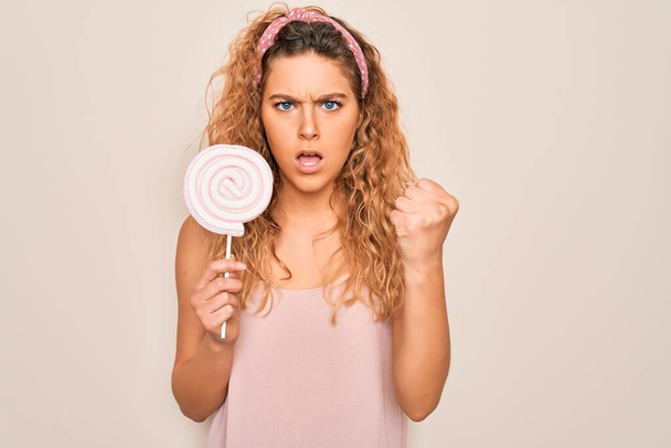 Giovane bella donna bionda con gli occhi blu mangiare caramelle dolci lecca-lecca su sfondo rosa infastidito e frustrato gridando con rabbia, pazzo e urlando con mano sollevata, concetto di rabbia
 - Foto, immagini