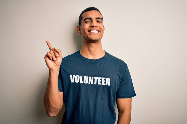 Mladý pohledný afroameričan se dobrovolně hlásí v tričku s dobrovolnickým vzkazem s širokým úsměvem na tváři, ukazuje rukou a prstem na stranu a dívá se do kamery. - Fotografie, Obrázek