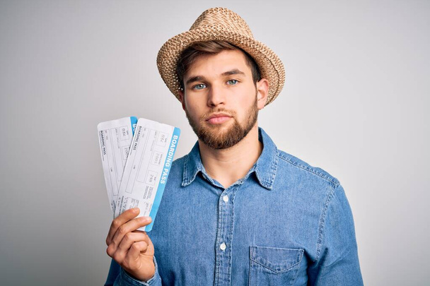 Joven turista rubio con ojos azules en vacaciones con sombrero sosteniendo tarjeta de embarque con una expresión segura en la cara inteligente pensando en serio
 - Foto, Imagen