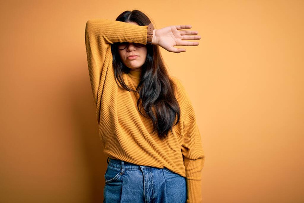 Young brunette woman wearing glasses and casual sweater over yellow isolated background covering eyes with arm, looking serious and sad. Sightless, hiding and rejection concept - Photo, Image