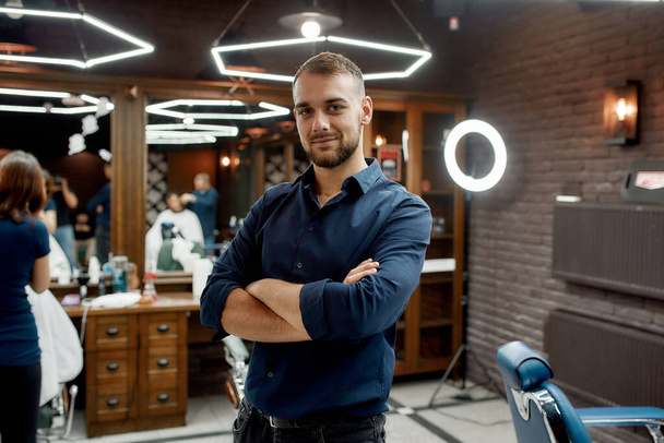 Working day at barbershop. Young and handsome barber in shirt looking at camera while standing at modern barbershop - Фото, изображение