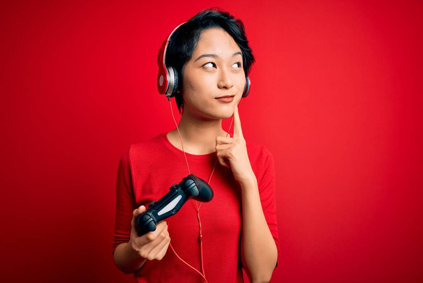 Jovem bonita asiático gamer menina jogar vídeo game usando joystick e fones de ouvido sério rosto pensando em questão, ideia muito confusa
 - Foto, Imagem