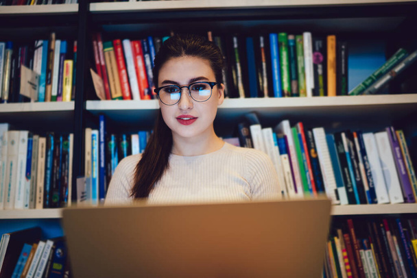 Low angle of dark haired focused woman in glasses sitting in library next to bookcase with colorful books and thoughtfully using laptop - Foto, afbeelding