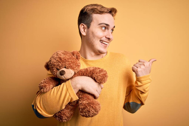 Молодий красивий рудий чоловік тримає плюшевий ведмідь тваринну іграшку над жовтим тлом, спрямовуючи великий палець в сторону посміхаючись з відкритим ротом
 - Фото, зображення