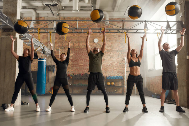 Enhance your Health. Full length shot of sportive people in black sportswear using exercise ball while having workout at industrial gym. Group training, teamwork concept - Fotografie, Obrázek