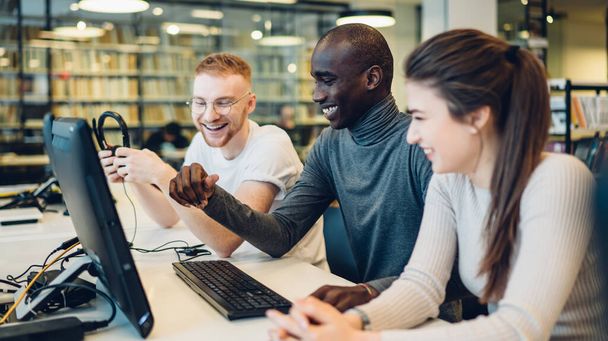 Happy multiethnic young people laughing and looking at screen while browsing computer with headphones and sitting at desk in modern library - 写真・画像