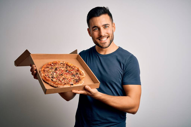 Joven hombre guapo sosteniendo la caja de entrega con deliciosa pizza italiana sobre fondo blanco con una cara feliz de pie y sonriendo con una sonrisa confiada mostrando los dientes
 - Foto, imagen