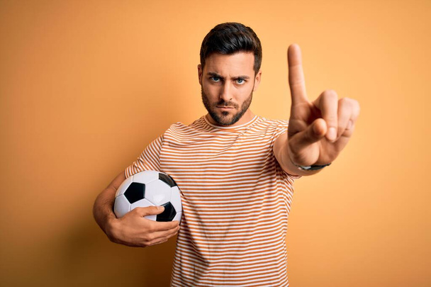 Handsome player man with beard playing soccer holding footballl ball over yellow background Pointing with finger up and angry expression, showing no gesture - Foto, afbeelding