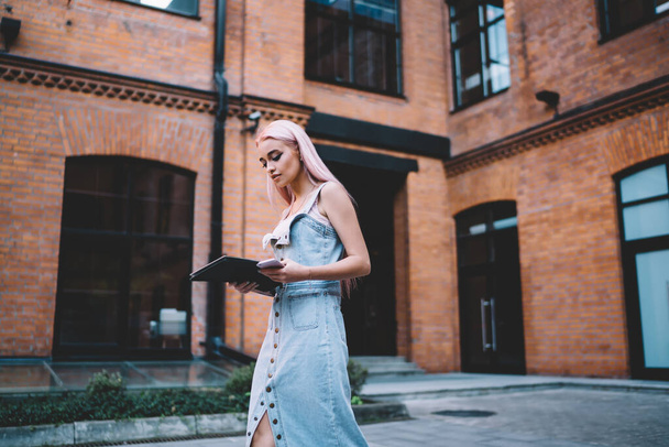 Slim blonde young female with long hair in denim sleeveless long dress carrying black folder and phone walking in street along building of red brick  - Фото, зображення
