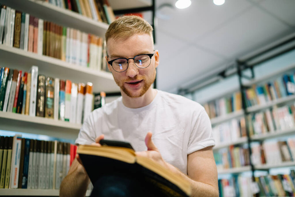 D'en bas coûteux sourire homme barbu adulte en chemise blanche et des lunettes avec livre ouvert sur les genoux en utilisant le téléphone et en se concentrant sur l'écran dans la bibliothèque moderne lumière - Photo, image