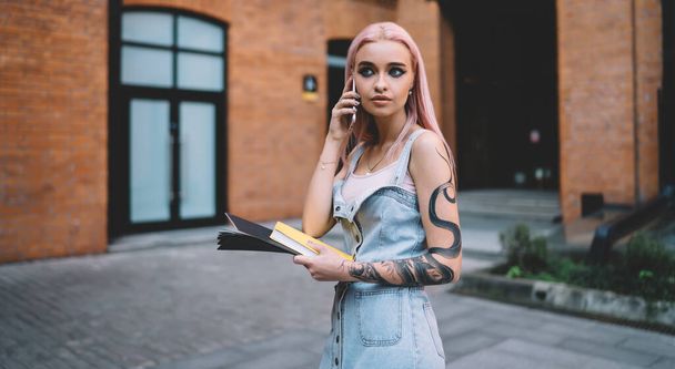 Fashionable lady with tattoo and long pink hair dressed in jeans dress talking on mobile phone holding yellow book in hand - Foto, immagini