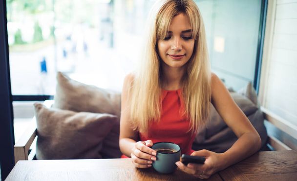 Young slightly smiling blonde female dressed in summer coral clothes browsing mobile phone while drinking coffee in cafe at daytime - Photo, Image