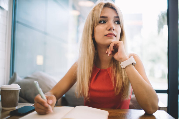 Young blond female in casual outfit looking away and thinking while sitting at table in cozy cafe and making notes in open planner - Photo, image