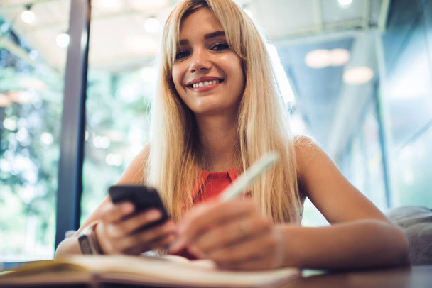 Low angle of glad young female with blond hair smiling and looking at camera while making notes and using smartphone at table in cafe - 写真・画像