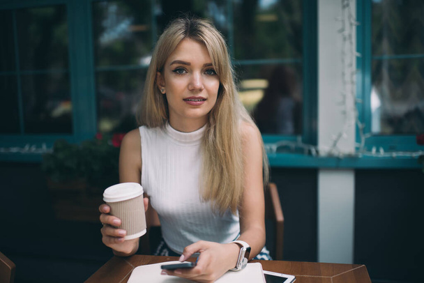 Young long haired blond woman using mobile phone while holding cup of coffee and sitting at table of coffee shop - Φωτογραφία, εικόνα
