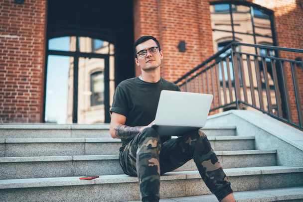 From below pensive adult male in khaki clothes looking at camera while sitting on stairs and interacting with laptop on street against entrance of blurred modern building with brown brick walls - Photo, Image