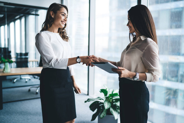 Side view of happy elegant ladies friendly smiling and confirming partnership with handshake while standing in modern office during working time - Φωτογραφία, εικόνα