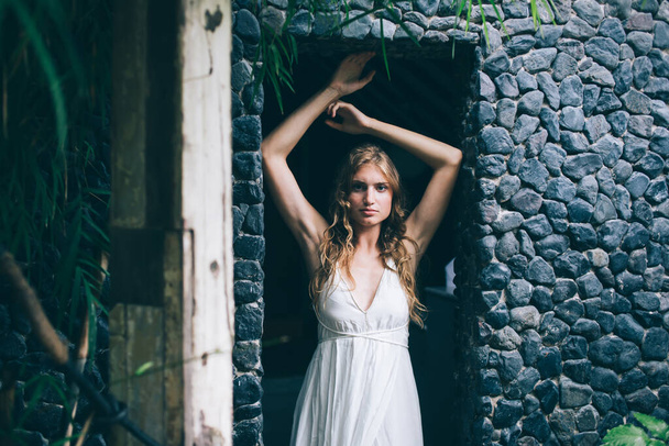 Charming thoughtful romantic female with light hair looking at camera while standing with raised hands in stony doorway on Bali - Foto, afbeelding