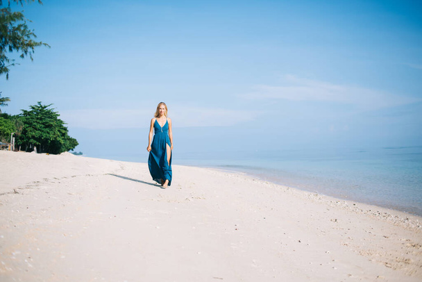 Young slender blonde female in flowing blue dress walking along beautiful sandy beach with clear blue seawater on sunny day with blue sky - Foto, Bild