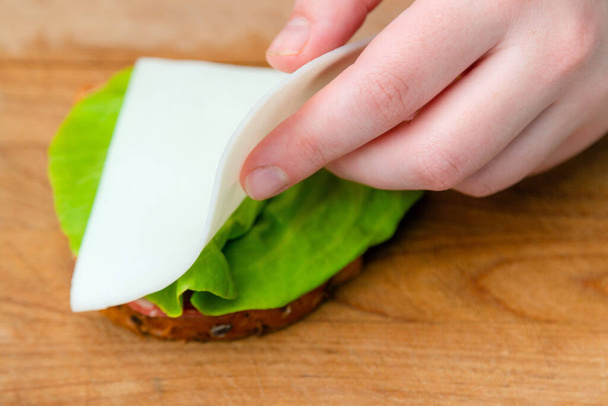 Close up of hand placing cheese on bread slice with salad leaf on wooden cutting board. Making sandwich - Foto, afbeelding