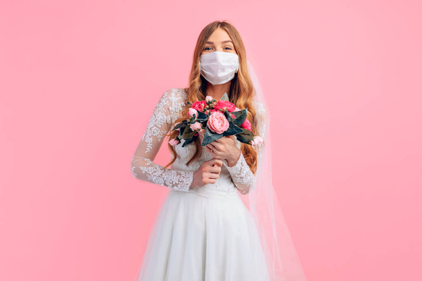 Beautiful bride in a wedding dress and medical protective mask on her face, on an isolated pink background. Wedding, quarantine, coronavirus - Fotó, kép