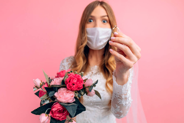 A bride in a wedding dress and a medical protective mask on her face shows her hand with an engagement ring on a pink background. Wedding, quarantine, coronavirus - Foto, immagini