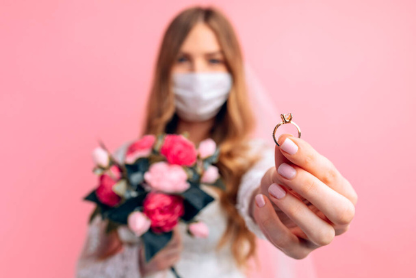 A bride in a wedding dress and a medical protective mask on her face shows her hand with an engagement ring on a pink background. Wedding, quarantine, coronavirus - Fotoğraf, Görsel