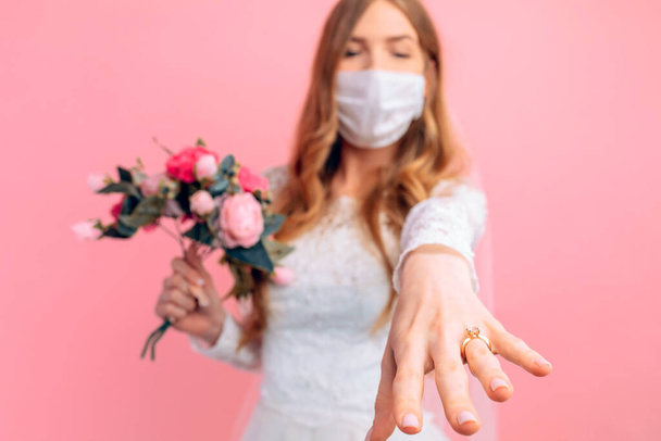 A bride in a wedding dress and a medical protective mask on her face shows her hand with an engagement ring on a pink background. Wedding, quarantine, coronavirus - Photo, image