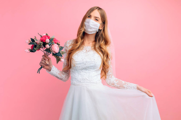Portrait of a beautiful bride with a wedding bouquet in a medical protective mask on her face. Blonde with curly hair on a pink background. Quarantine, wedding, coronavirus - Fotoğraf, Görsel