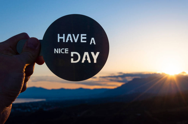 Рука держит металлический диск с надписью "Хорошего дня" вырезанный, размытый фон с горами и побережьем моря до восхода солнца
 - Фото, изображение