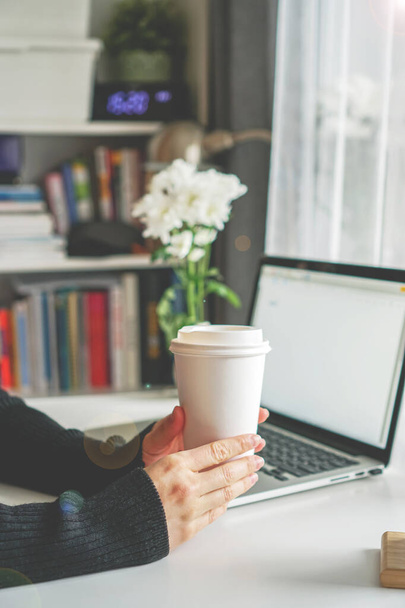 Bir kız masada oturuyor ve elinde bir bardak kahve tutuyor. Yanında bir dizüstü bilgisayar duruyor. - Fotoğraf, Görsel