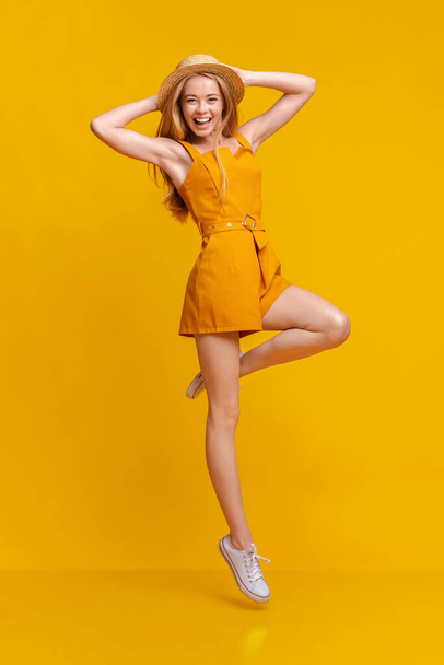 Summer Fashion. Joyful Girl In Jumpsuit And Hat Jumping On Yellow Background - Фото, изображение