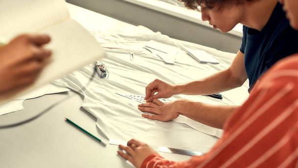 Production process. Male hands working with sketches or patterns at studio and preparing fabric. Group of millennials creating clothes together - Foto, afbeelding