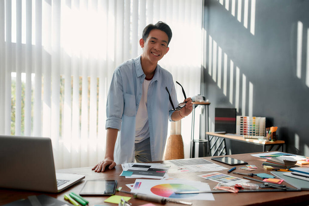 Happy at work. Cheerful asian man holding eyeglasses and smiling at camera while leaning on a table with a lot of creative stuff on it. Young designer in casual wear working in the modern office - Foto, imagen