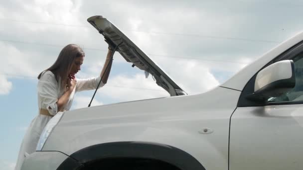 vrouw drukt verbijstering wanneer ze kijkt onder de motorkap van de auto, problemen met haar kapotte auto, het openen van de motorkap en bellen voor hulp op haar mobiele telefoon - Video