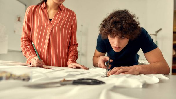 Born to create. Young male designer cutting white fabric textile in a studio. Group of creative millennials working together - 写真・画像