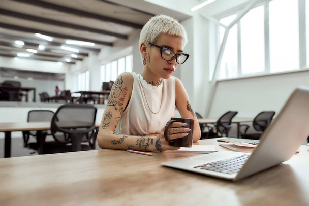 Thinking about business. Young thoughtful tattooed business woman in eyewear holding a cup and looking at laptop screen while sitting alone in the modern office. - Fotó, kép