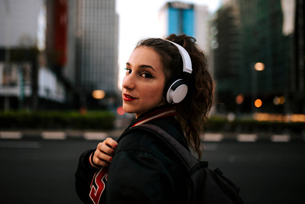 Belle femme espagnole écoutant de la musique avec des écouteurs et un téléphone intelligent à l'extérieur. Fille latine en vêtements de sport avec coiffure bouclée en arrière-plan urbain
. - Photo, image