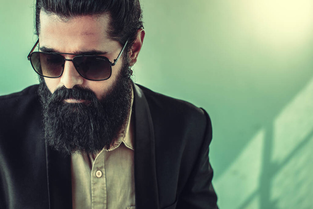 front view of handsome bearded man with a man bun hairstyle posing in studio wearing a suit and sunglasses on green background, male model isolated fashion portrait photograph. - Photo, Image