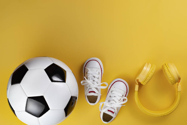 Close-up photo of yellow headphones, white sneakers and traditional style soccer ball over yellow background. - Foto, Bild