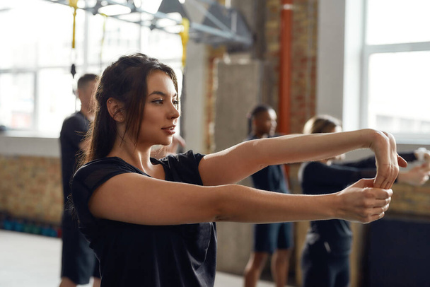 Make yourself proud. Portrait of young woman in black sportswear looking focused, stretching arms while working out in industrial gym. Group training concept - Φωτογραφία, εικόνα