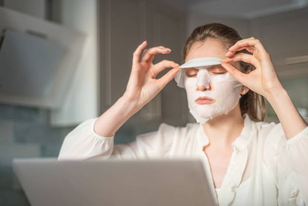 Finish the care procedure to rejuvenate the skin of the face. Young girl removes a cloth mask for the face, working at home for a laptop - Foto, immagini