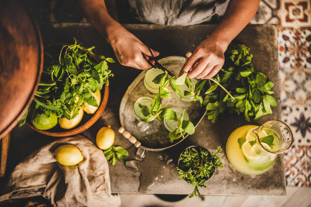 Hands of woman making fresh homemade lemonade. Woman decorating cold citrus lemonade in glasses with fresh mint leaves over grey marble kitchen counter and oriental tile floor, top view - Foto, Bild