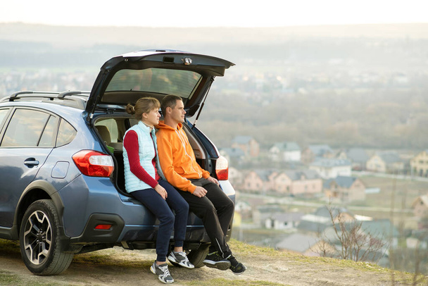 Happy couple standing together near a car with open trunk enjoying view of rural landscape nature. Man and woman leaning on family vehicle luggage compartment. Weekend travel and holidays concept. - Foto, Imagen