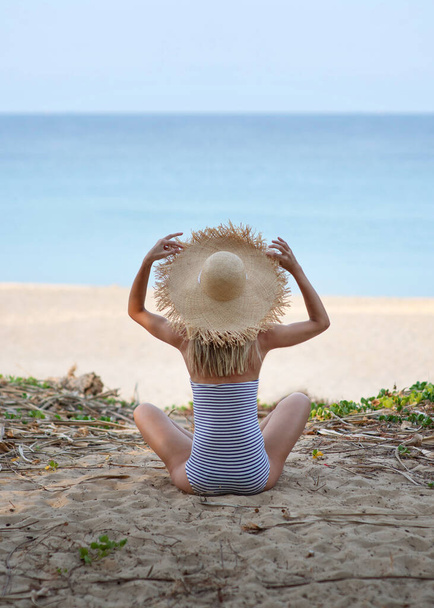 Girl in a straw hat sits and has a striped bathing suit on her exotic vacation. Tropoic island. Faceless. Tranquil rest on the beach near palms. The concept of rest, tourism and relaxation - Photo, Image