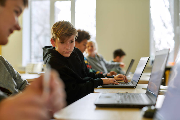 Szuka pomocy. Portret białego ucznia patrzącego na laptopa sąsiada, siedzącego w klasie z innymi uczniami podczas lekcji w nowoczesnej inteligentnej szkole - Zdjęcie, obraz